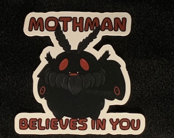 Puede incluir: Una pegatina con un personaje de dibujos animados de Mothman, con ojos rojos y un cuerpo negro. El texto "MOTHMAN" está encima del personaje, y "BELIEVES IN YOU" debajo, todo en un esquema de color rojo y blanco.