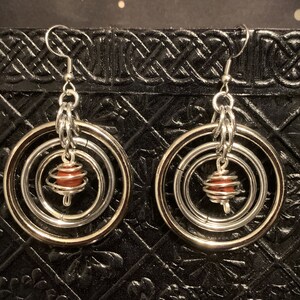 May include: Silver hoop earrings with a dangling red bead in a wire cage. The earrings have three hoops, one inside the other.