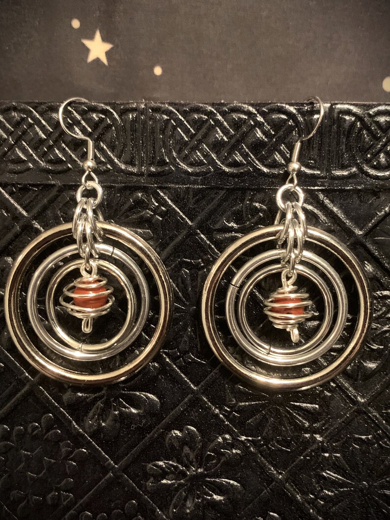 May include: Silver hoop earrings with a dangling red bead in a wire cage. The earrings have three hoops, one inside the other.