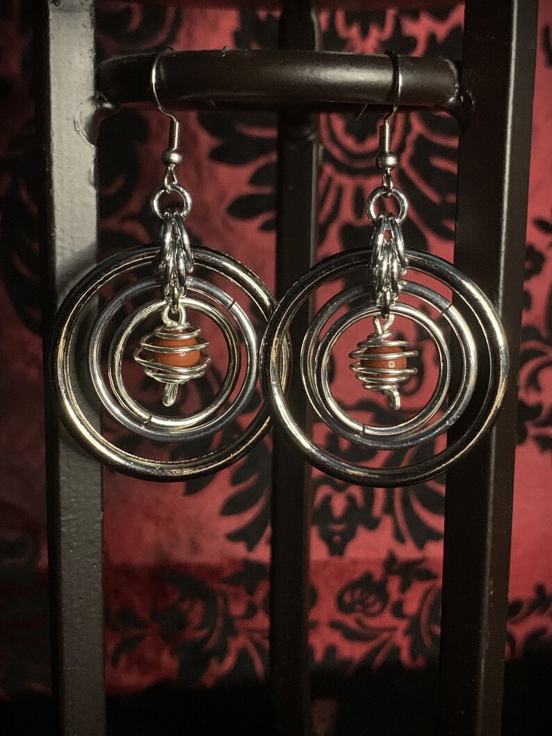 May include: Silver hoop earrings with multiple rings and a small orange bead in the center.