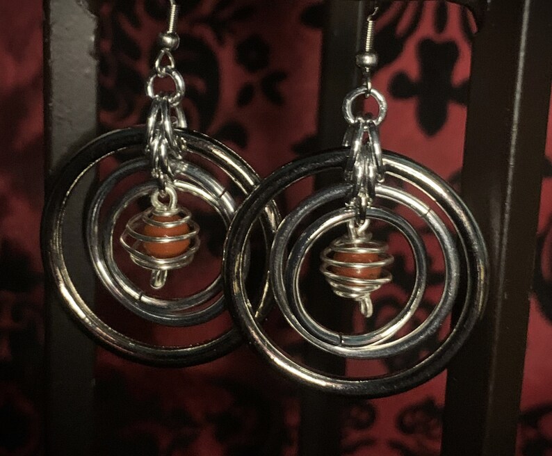 May include: Silver hoop earrings with three concentric circles and a red gemstone encased in a wire cage hanging from the center.