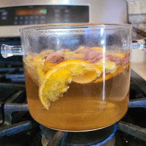 Peut inclure: Une casserole en verre avec des zestes d'orange et un liquide brun est sur une cuisinière avec une flamme bleue.