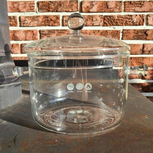 Peut inclure: Une casserole en verre transparent avec un couvercle et deux poignées. La casserole est marquée sur le côté pour indiquer qu'elle peut être utilisée sur différents types de plaques de cuisson.