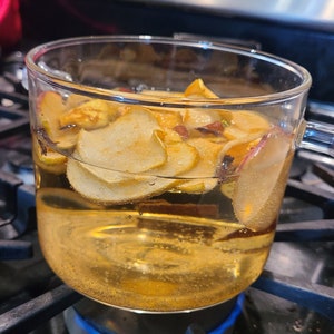 May include: A glass jar filled with apple slices and a clear liquid is sitting on a stovetop burner with a blue flame. The jar is on a black metal burner grate.