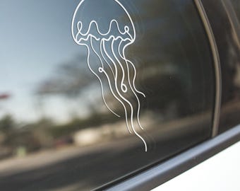 May include: A white vinyl decal of a jellyfish is affixed to a car window. The decal features a minimalist line drawing of the marine animal, with a bell-shaped body and flowing tentacles. The car window reflects the surrounding environment.