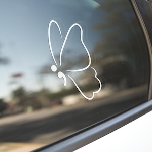May include: A white butterfly decal is affixed to a car window. The decal is a simple outline of a butterfly, with its wings and body clearly defined. The car window reflects the outside environment.