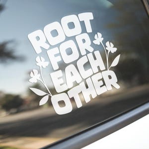 May include: White vinyl decal with the words "ROOT FOR EACH OTHER" in a bold, rounded font, accented with floral illustrations. The decal is applied to a car window, with a blurred outdoor background visible.
