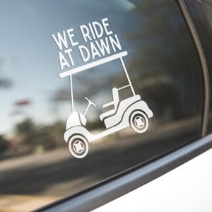 May include: White vinyl decal of a golf cart with the text "WE RIDE AT DAWN". The golf cart is depicted in a simple, clean style, with visible wheels, a steering wheel, and a seat. The decal is applied to a car window.