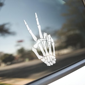 May include: A white skeleton hand making the peace sign. The hand is on a car window.