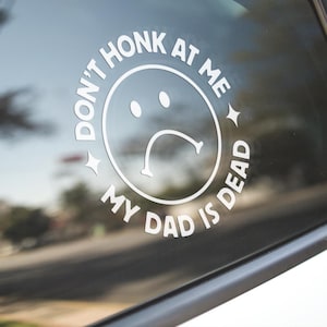 May include: White vinyl car decal with the text "DON'T HONK AT ME MY DAD IS DEAD" surrounding a sad face emoji. The decal is applied to a car window, with a blurred background.