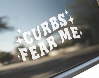 May include: White vinyl decal with the text "Curbs Fear Me" in a retro font with sparkling stars on a car window.