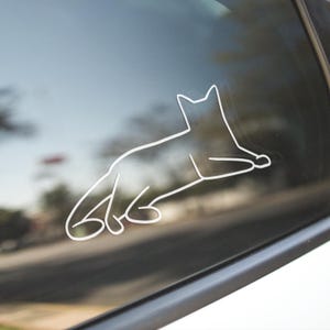 May include: White outline of a cat in a relaxed pose, applied to a car window. The minimalist design features the cat's silhouette, with details of its ears, legs, and tail. The sticker is a simple, elegant decoration.