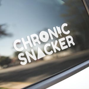 May include: White vinyl sticker with the text "CHRONIC SNACKER" in a bold, sans-serif font. The design incorporates a star, a triangle, and a knife icon within the lettering. The sticker is affixed to a car window, with a blurred outdoor scene visible.