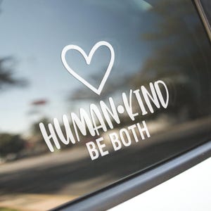 May include: A white vinyl decal with a heart and the text "HUMAN-KIND BE BOTH" on a black car window.