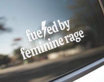 Op de afbeelding: Witte vinyl sticker met de tekst "fueled by feminine rage" en een blikseminslag. De sticker is aangebracht op een autoruit, met een vage achtergrond van een straat en bomen.