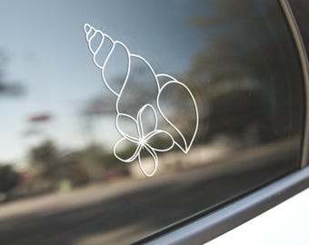 Puede incluir: Calcomanía de vinilo blanco con un diseño de concha, flor y hoja. La calcomanía está aplicada a la ventana de un coche, con el contorno del diseño visible contra el cristal oscuro. La imagen está tomada al aire libre, con un fondo borroso de una calle.
