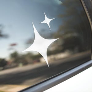 May include: A white car window with two white star stickers on it. The stars are in a layered design, with one star slightly overlapping the other.