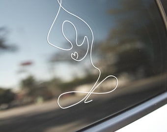 May include: White vinyl decal of a person in a yoga pose, applied to a car window. The design is a continuous line drawing, depicting a figure in a seated meditation position. The background shows a blurred outdoor scene.