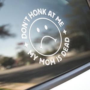 May include: White vinyl car decal with the text "DON'T HONK AT ME MY MOM IS DEAD" encircling a sad face emoji. The decal is affixed to a car window, with a blurred background of trees and a road.