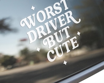 May include: White vinyl decal applied to a car window. The decal reads "WORST DRIVER BUT CUTE" in a stylized font, with small star accents. The background shows a blurred view of trees and a road.