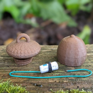 May include: Two brown 3D printed acorn-shaped containers, one with a loop handle, and a rolled up piece of paper with the text "Geocaching Game Piece" on it.
