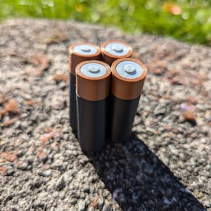 May include: Four brown and black AA batteries on a gray, textured surface.