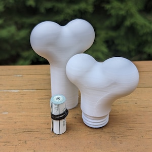 Pode incluir: Dois objetos brancos em forma de osso impressos em 3D, um maior que o outro, sobre uma superfície de madeira. Um pequeno pedaço de papel enrolado com fita adesiva preta fica na frente dos objetos.