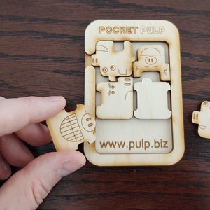 Könnte beinhalten: Holzpuzzle mit dem Text "POCKET PULP" und "www.pulp.biz". Das Puzzle hat einen rechteckigen Rahmen und schweineförmige Teile. Ein Teil wird von einer Hand gehalten. Das Puzzle ist aus hellem Holz gefertigt.