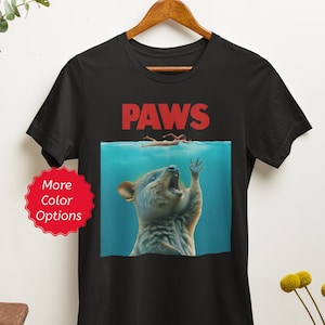 Puede incluir: Camiseta negra con un dise&ntilde;o parodia que presenta la palabra "PAWS" en rojo, sobre una ilustraci&oacute;n de un quokka en el agua, extendiendo la mano hacia un nadador. El dise&ntilde;o es una versi&oacute;n humor&iacute;stica de un cartel de pel&iacute;cula cl&aacute;sico.