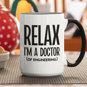 May include: White ceramic mug with black handle and rim. The mug has the text "RELAX I'M A DOCTOR (OF ENGINEERING)" in black, bold letters. A red and white polka-dotted container is in the background.