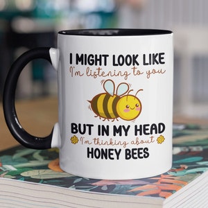 Puede incluir: Taza de café de cerámica blanca con asa negra y borde negro. La taza tiene una abeja de dibujos animados y el texto "Puede que parezca que te estoy escuchando, pero en mi cabeza estoy pensando en abejas."