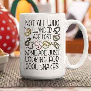 May include: White ceramic mug with a black handle. The mug displays the text "NOT ALL WHO WANDER ARE LOST, SOME ARE JUST LOOKING FOR COOL SNAKES" and illustrations of colourful snakes.