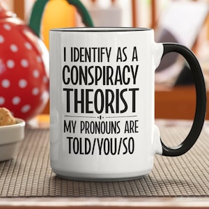 May include: White coffee mug with a black handle and black rim. The mug has black text that reads "I identify as a conspiracy theorist. My pronouns are told/you/so."