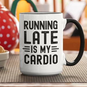 Könnte beinhalten: Weiße Keramik-Kaffeetasse mit schwarzem Henkel. Die Tasse hat schwarzen Text mit der Aufschrift "RUNNING LATE IS MY CARDIO".