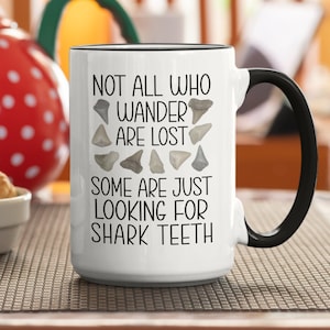 May include: White ceramic mug with a black handle. The mug has a black rim and features the text "Not all who wander are lost. Some are just looking for shark teeth." with a graphic of shark teeth.
