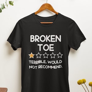May include: Black t-shirt with the text "BROKEN TOE" in white, along with a rating of one out of five stars and the words "TERRIBLE. WOULD NOT RECOMMEND." The shirt is hanging on a wooden hanger.