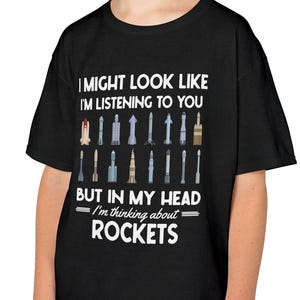 May include: Black t-shirt with the text "I MIGHT LOOK LIKE I'M LISTENING TO YOU BUT IN MY HEAD - I'm thinking about ROCKETS." The shirt features a series of rocket illustrations in various colors and designs.