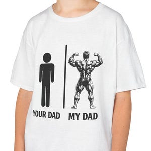 Peut inclure: T-shirt blanc avec un graphique comparant un bonhomme allumette étiqueté "YOUR DAD" à une figure musclée étiquetée "MY DAD". Le t-shirt est porté par un jeune.