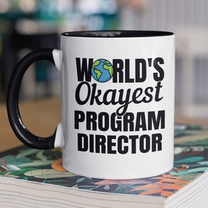 Peut inclure: Une tasse en céramique blanche avec un bord et une poignée noirs. La tasse porte une inscription noire qui dit "WORLD'S Okayest PROGRAM DIRECTOR" avec une petite image de la Terre en dessin animé.
