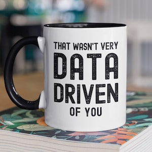以下が含まれることがあります： 黒色の取っ手と縁取りが付いた白いコーヒーカップ。カップには黒い文字で「That wasn't very data driven of you.」と書かれています。