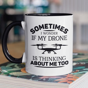 Op de afbeelding: Witte keramische mok met een zwarte rand en handvat. De mok heeft een zwarte afbeelding met de tekst "Sometimes I wonder if my drone is thinking about me too." met een silhouet van een drone.