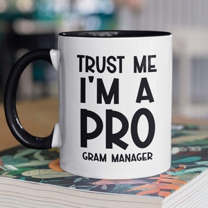 May include: White ceramic mug with a black rim and handle. The mug has black text that reads "Trust me I'm a Pro Gram Manager". The mug is sitting on a book with a colorful floral pattern.