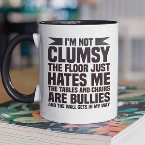 Peut inclure: Une tasse en céramique blanche avec un bord et une poignée noirs. La tasse a un texte noir qui dit "Je ne suis pas maladroit, le sol me déteste, les tables et les chaises sont des brutes et le mur me gêne."