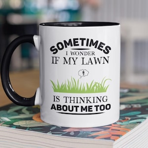 May include: White ceramic mug with a black interior and handle. The mug features the text "SOMETIMES I WONDER IF MY LAWN IS THINKING ABOUT ME TOO" in black and green. The mug is sitting on a book.