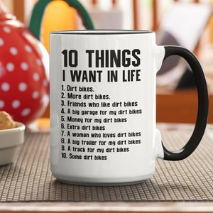 Könnte beinhalten: Eine weiße Keramiktasse mit schwarzem Henkel. Die Tasse hat einen schwarzen Rand und schwarzen Text, der "10 THINGS I WANT IN LIFE" lautet, gefolgt von einer nummerierten Liste von Artikeln, die mit Dirtbikes zu tun haben.