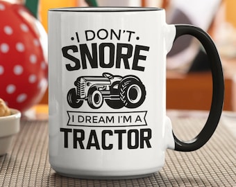 Könnte beinhalten: Eine weiße Keramik-Kaffeetasse mit schwarzem Henkel. Die Tasse zeigt eine Schwarzweiß-Illustration eines Traktors und den Text "Ich schnarche nicht, ich träume, ein Traktor zu sein."
