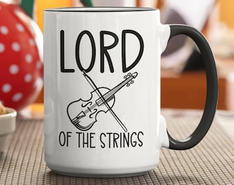 May include: White ceramic coffee mug with a black handle. The mug has a black and white illustration of a violin and the text "LORD OF THE STRINGS".