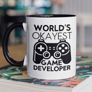 Könnte beinhalten: Weiße Kaffeetasse mit schwarzem Rand und Henkel. Die Tasse zeigt eine schwarz-weiße Grafik eines Videospiel-Controllers und den Text "WORLD'S OKAYEST GAME DEVELOPER".