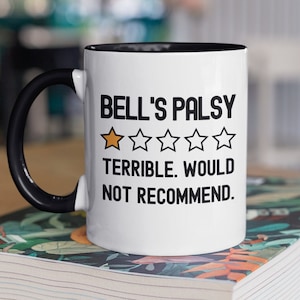 May include: White coffee mug with a black handle and black rim. The mug has black text that reads "Bell's Palsy" with a one-star rating and the text "Terrible. Would not recommend."