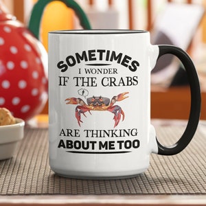 Op de afbeelding: Een witte keramische mok met een zwarte rand en handvat. De mok heeft een zwart-witte afbeelding van een krab met de tekst "Sometimes I wonder if the crabs are thinking about me too."